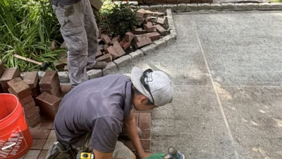 Two crew members installing brick pavers on a patio with a wheelbarrow and ornamental grasses nearby