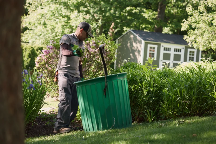 A Lawnscapes crew member in a gray uniform and cap trimming flowering shrubs into a green waste bin in a well-maintained garden bed, with a storage shed and lush greenery in the background.