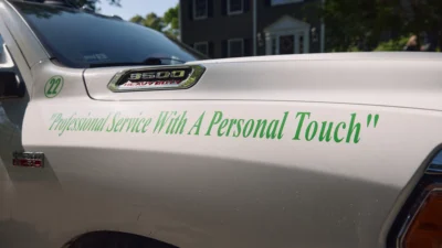Close-up of a white Lawnscapes work truck door with green script reading "Professional Service With A Personal Touch," parked in front of a residential home.