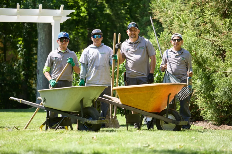 Four Lawnscapes crew members in gray uniforms posing with wheelbarrows, rakes, and shovels on a residential lawn, with a white pergola and evergreen trees in the background.